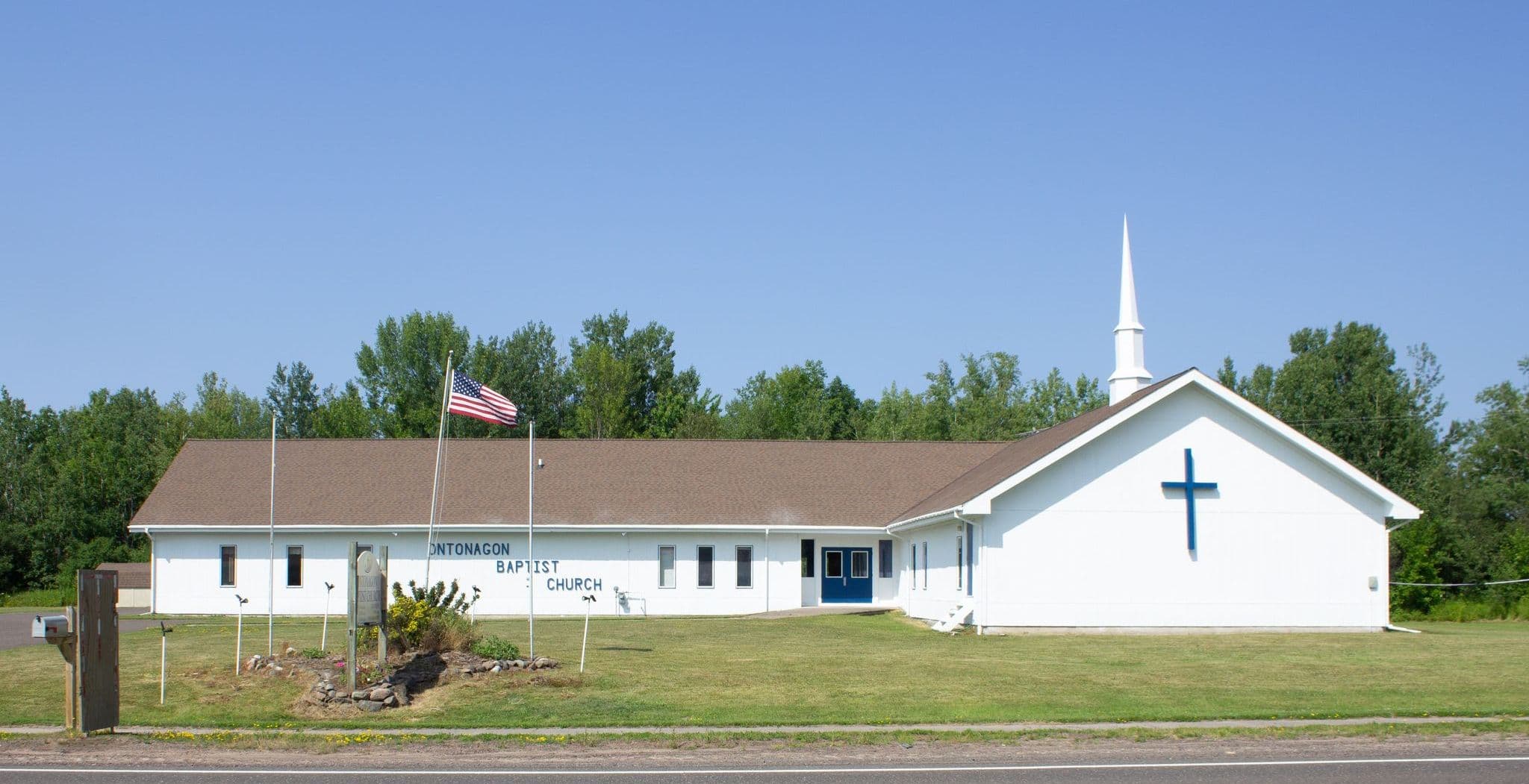 Ontonagon Baptist Church seen from Greenland Road — white siding, a brown roof, a white steeple and a blue cross on the gable, with the treeline behind and an open blue sky above